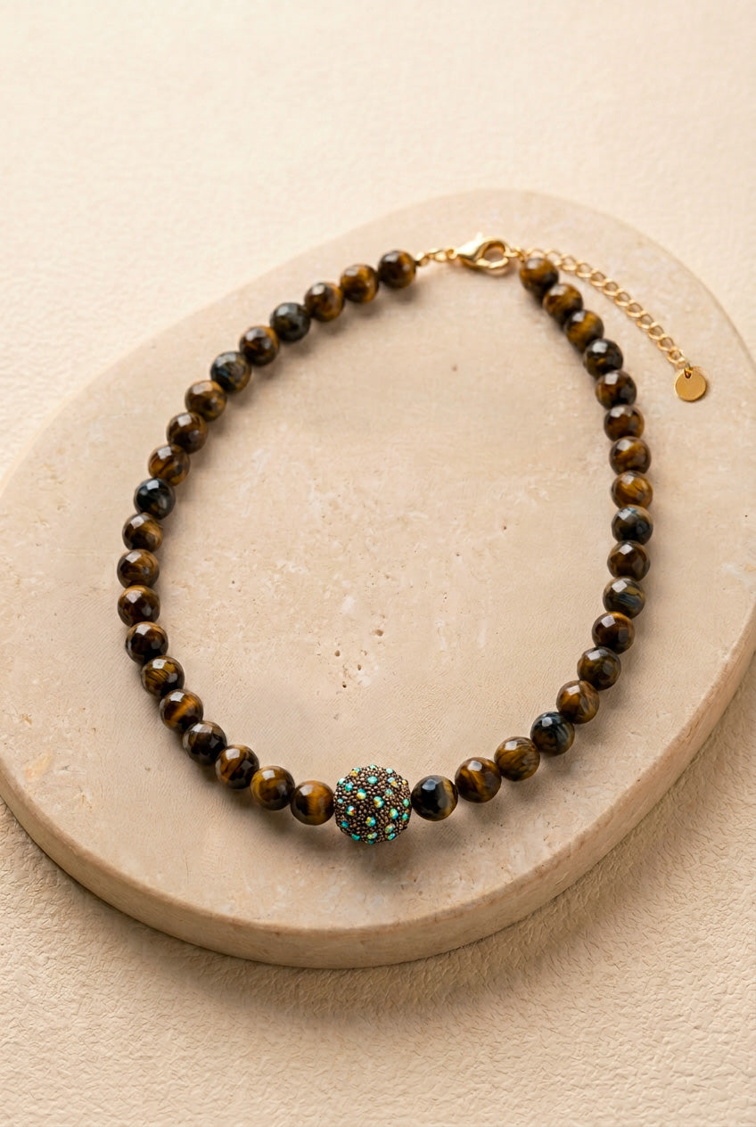 Vue d'ensemble du collier Gravy posé sur un support en pierre noire, montrant les perles facettées œil de tigre, la perle strass et le fermoir en acier inoxydable doré.