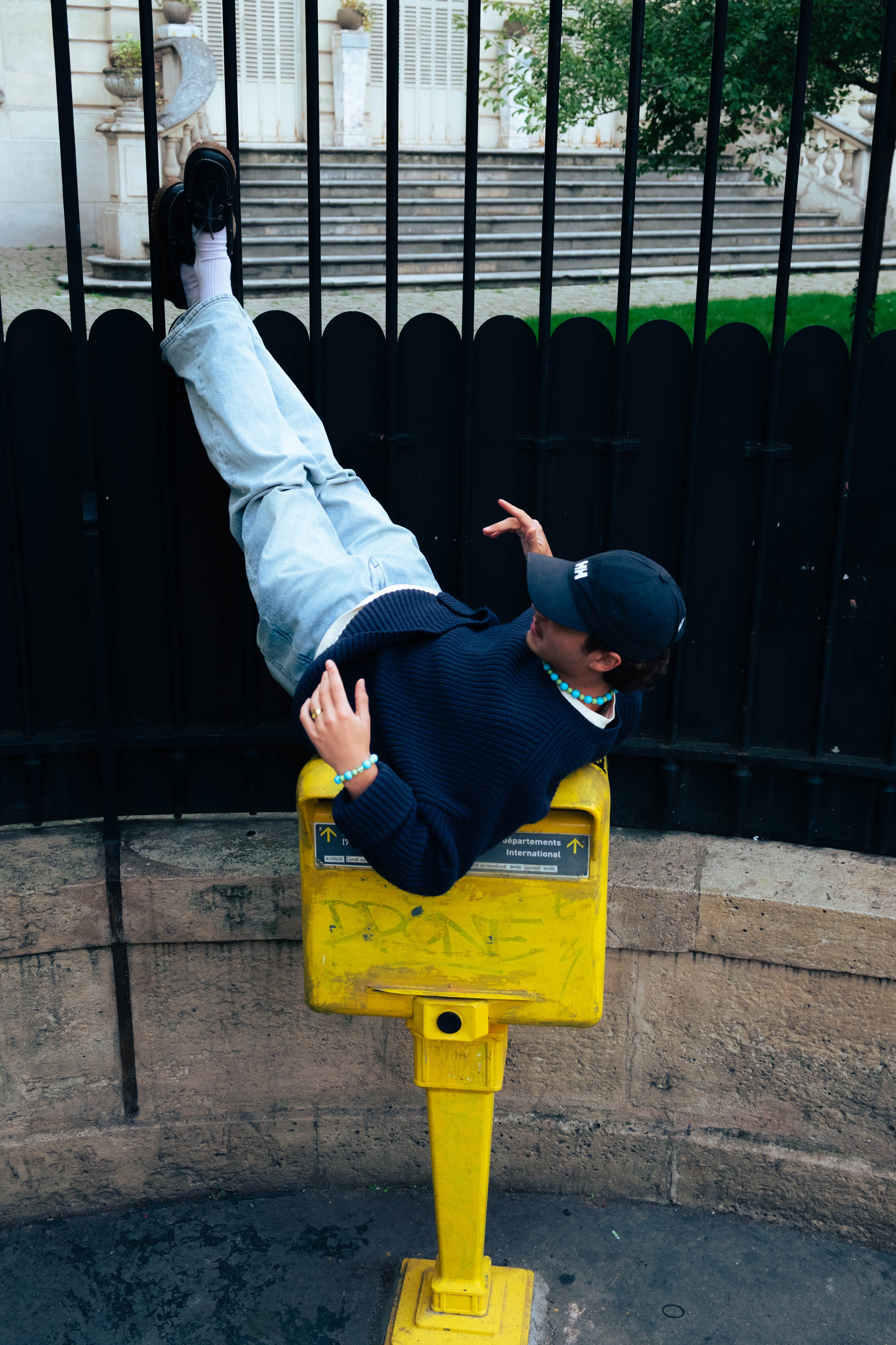 Homme portant un bracelet bleu Bourjeone, allongé sur une boîte aux lettres jaune à Paris.