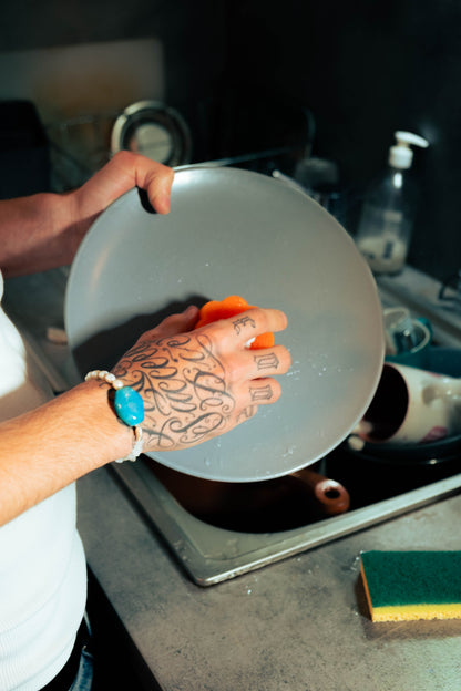 Main tatouée portant un bracelet turquoise Bourjeone en train de laver une assiette.