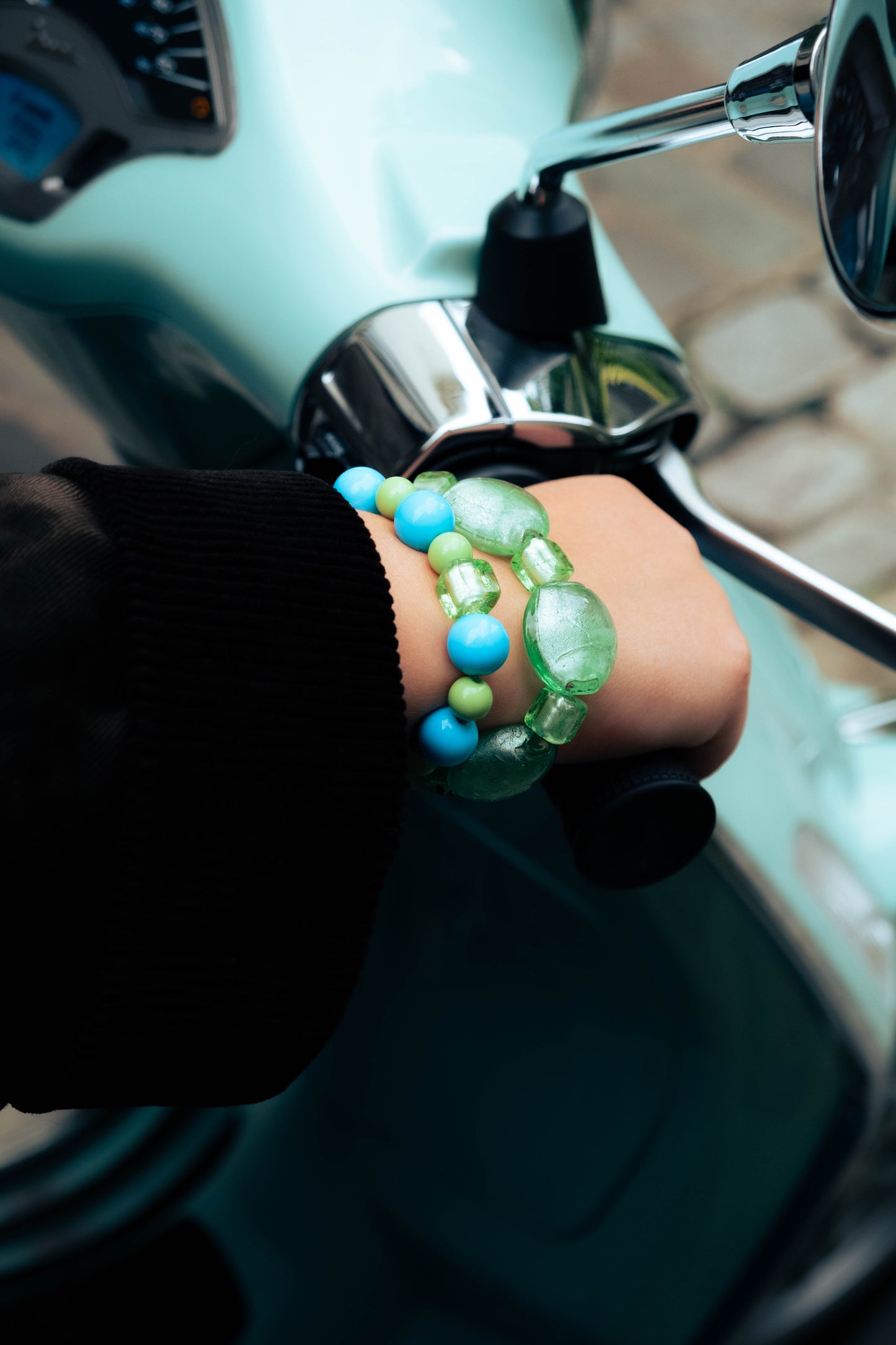 Main portant des bracelets Bourjeone bleus et verts sur un guidon de scooter mint à Paris.