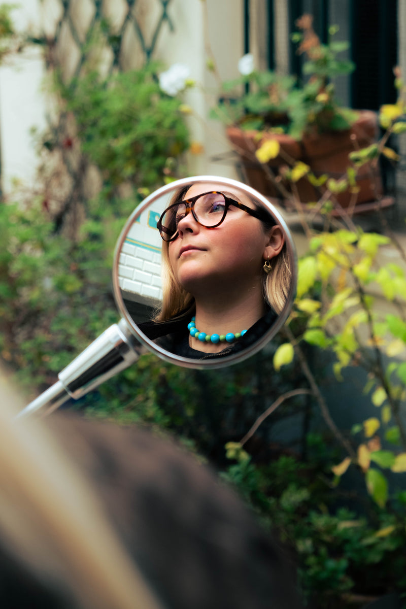 Reflet d’une femme portant un collier de perles bleues Bourjeone dans un rétroviseur.