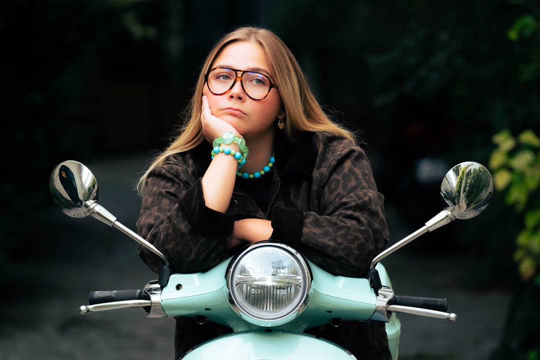 Femme appuyée sur un scooter mint portant un collier et des bracelets Bourjeone bleu et vert.