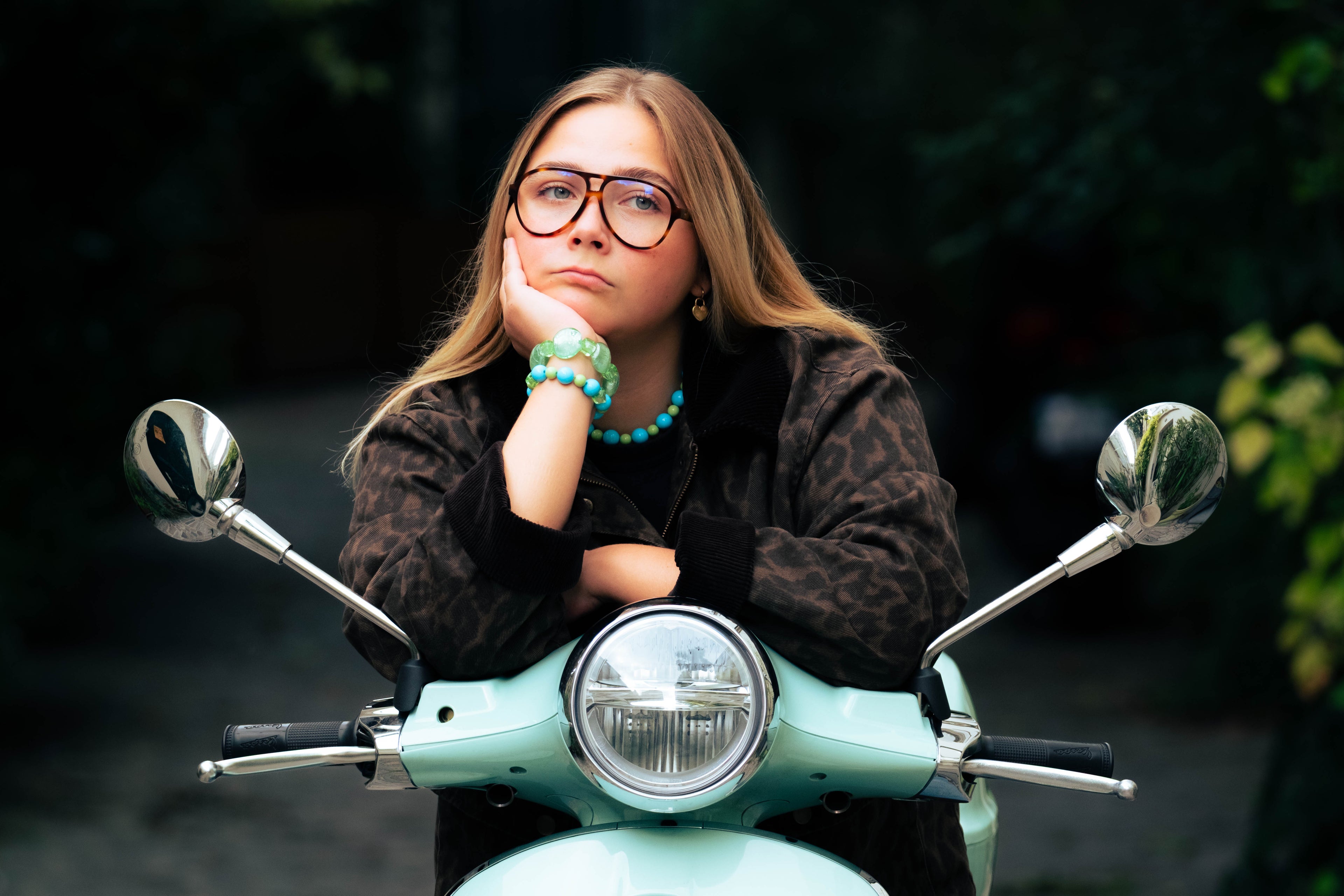 Femme appuyée sur un scooter mint portant un collier et des bracelets Bourjeone bleu et vert.
