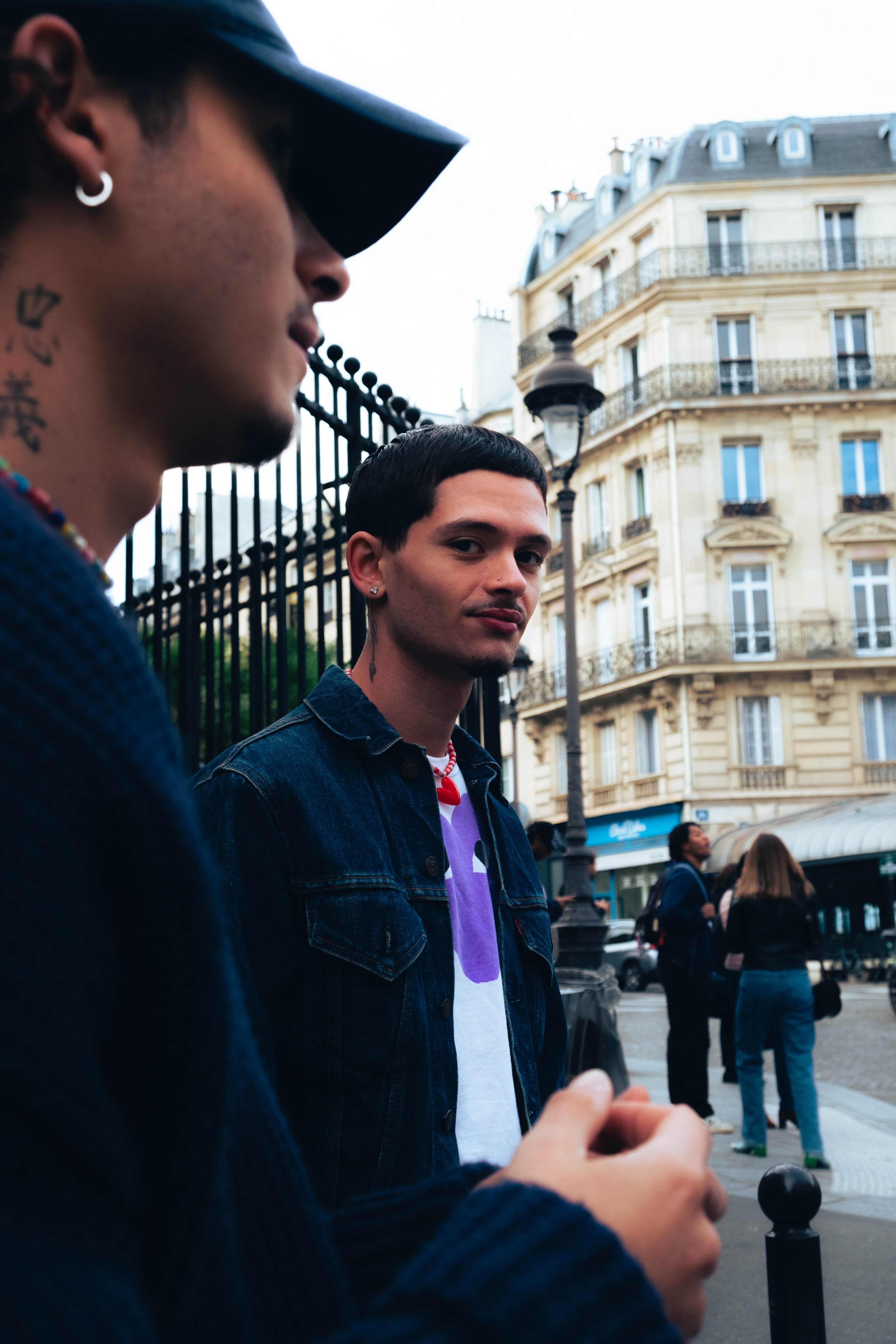 Homme portant un collier rouge Bourjeone dans une rue parisienne, face à un ami en pull bleu.