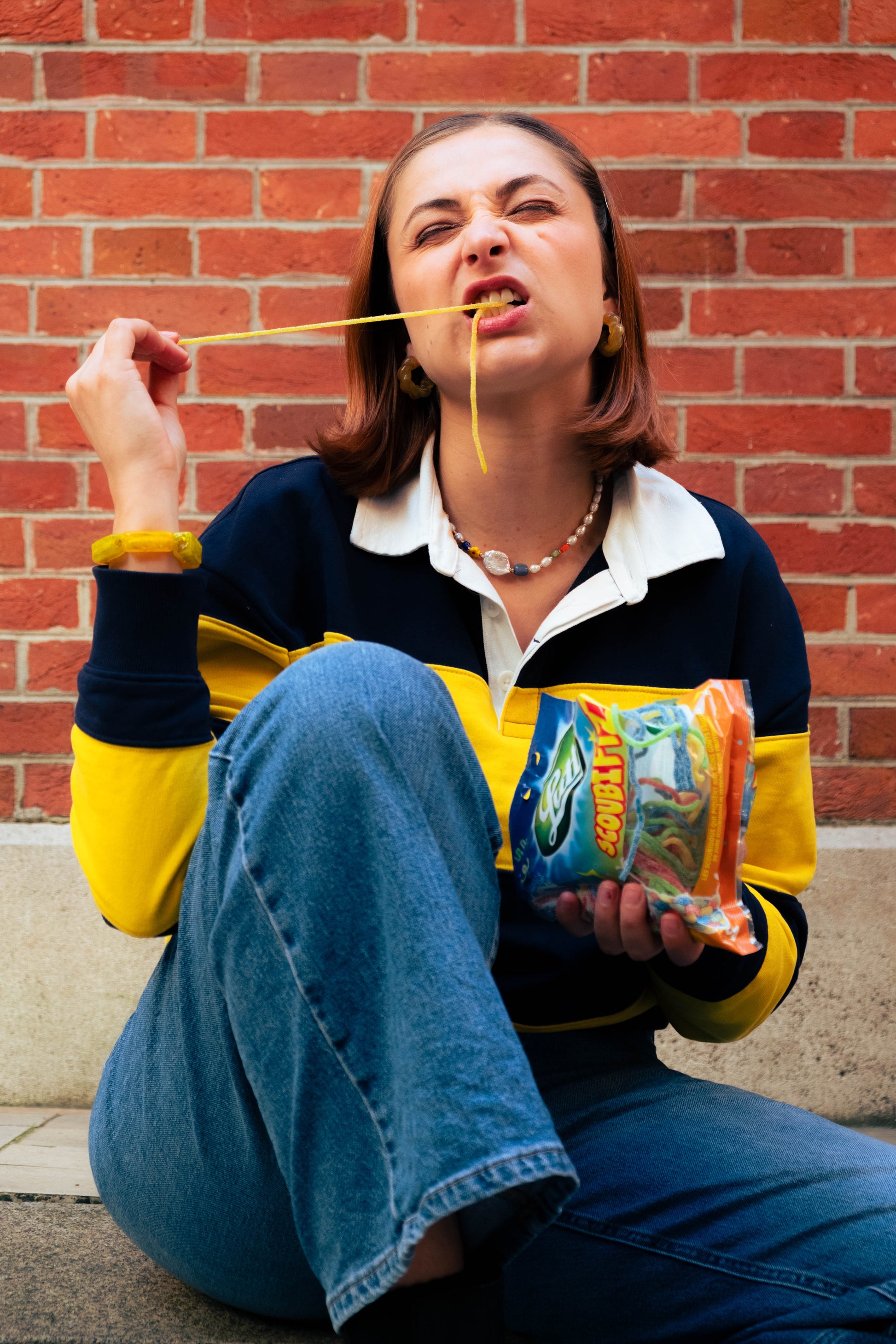 Femme portant un collier coloré Bourjeone et mangeant des bonbons acidulés devant un mur de briques.
