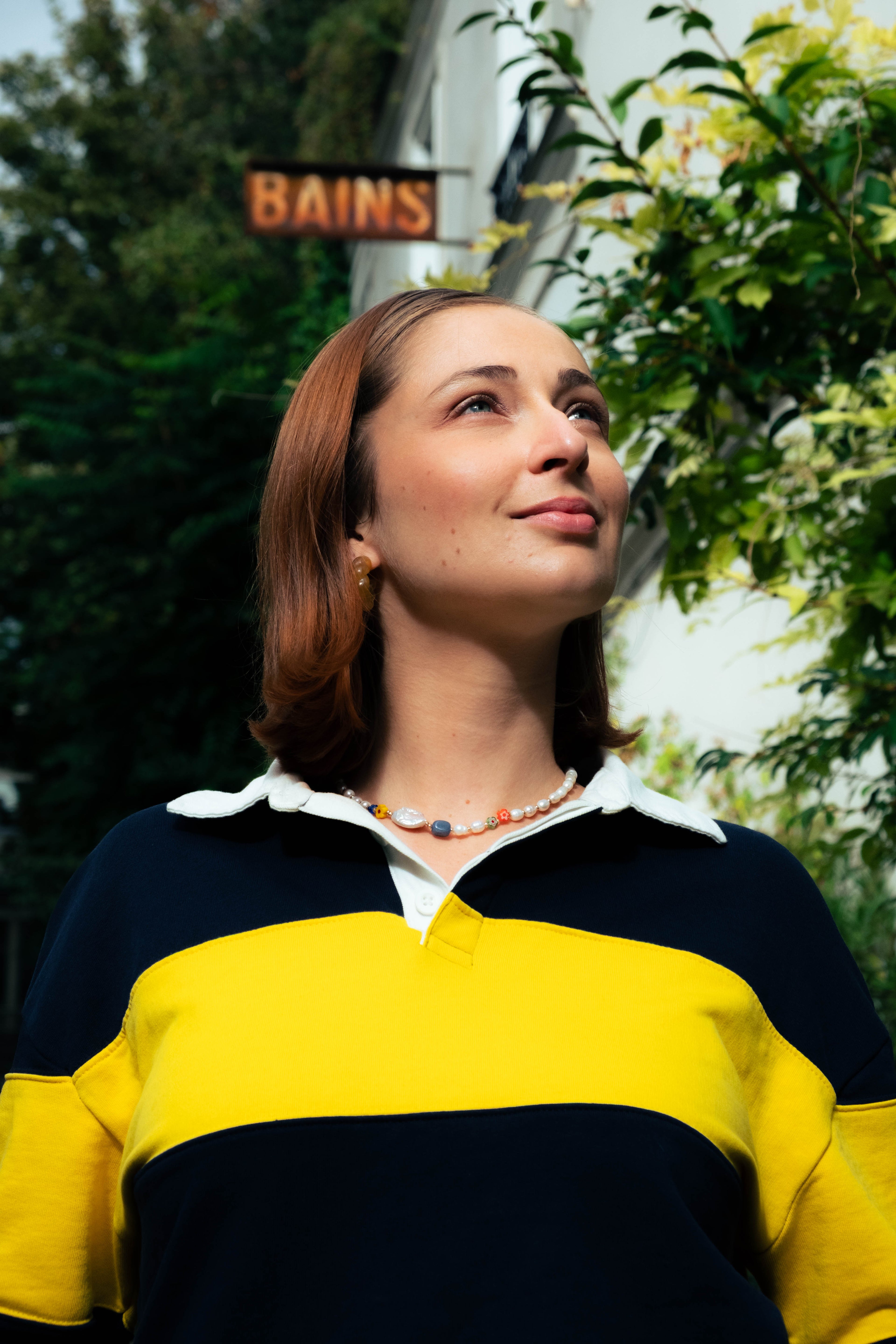 Femme portant un collier Bourjeone coloré, levant le regard vers le ciel dans une rue parisienne ensoleillée.