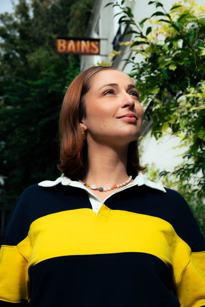 Femme portant un collier Bourjeone coloré, levant le regard vers le ciel dans une rue parisienne ensoleillée.