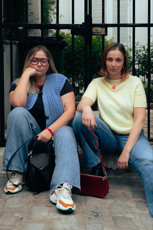 Deux femmes portant des colliers Bourjeone, assises sur un trottoir parisien devant une grille noire.