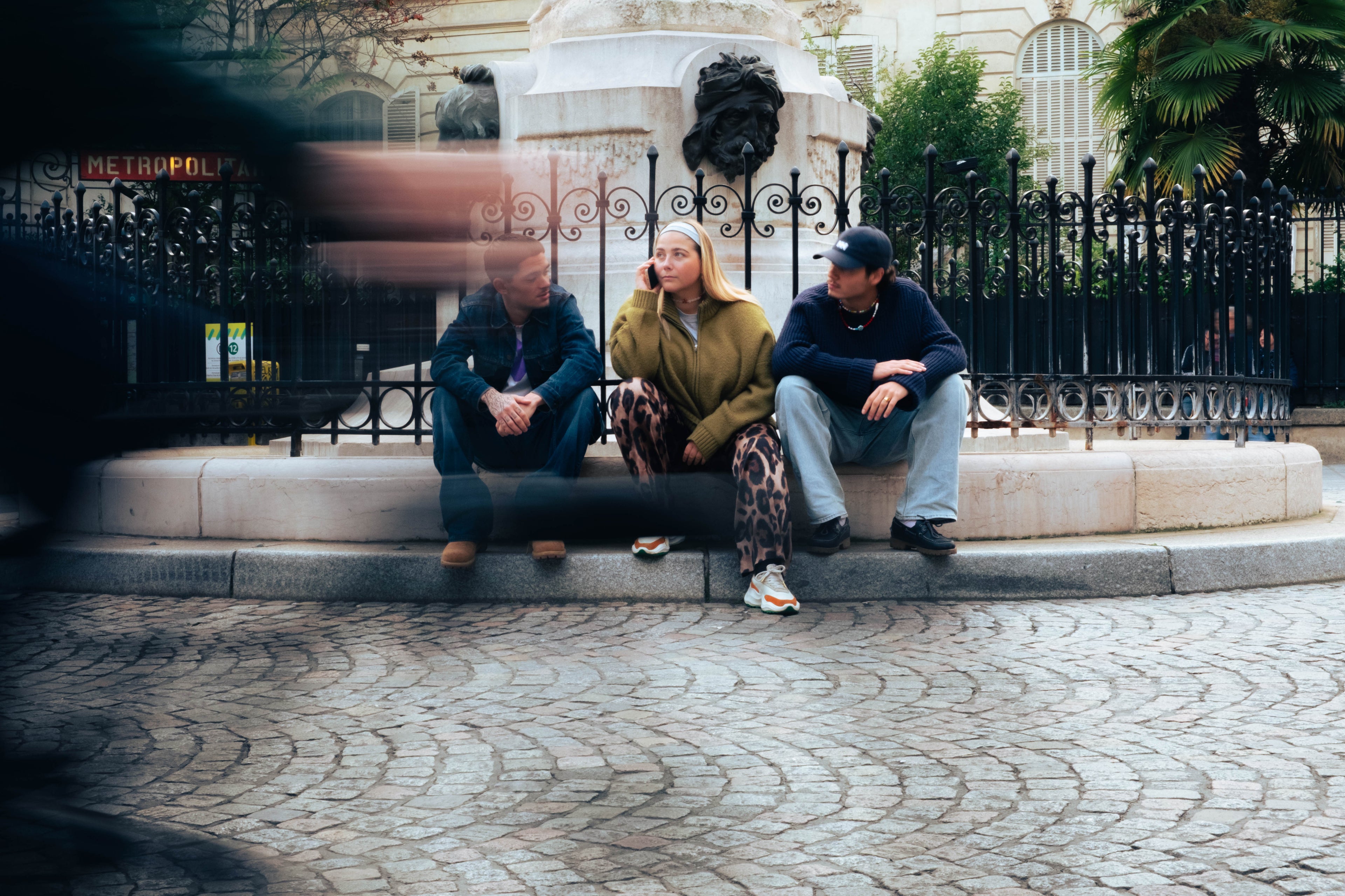 Trois jeunes assis sur une place parisienne portant des bijoux Bourjeone, avec un mouvement flou de vélos au premier plan.