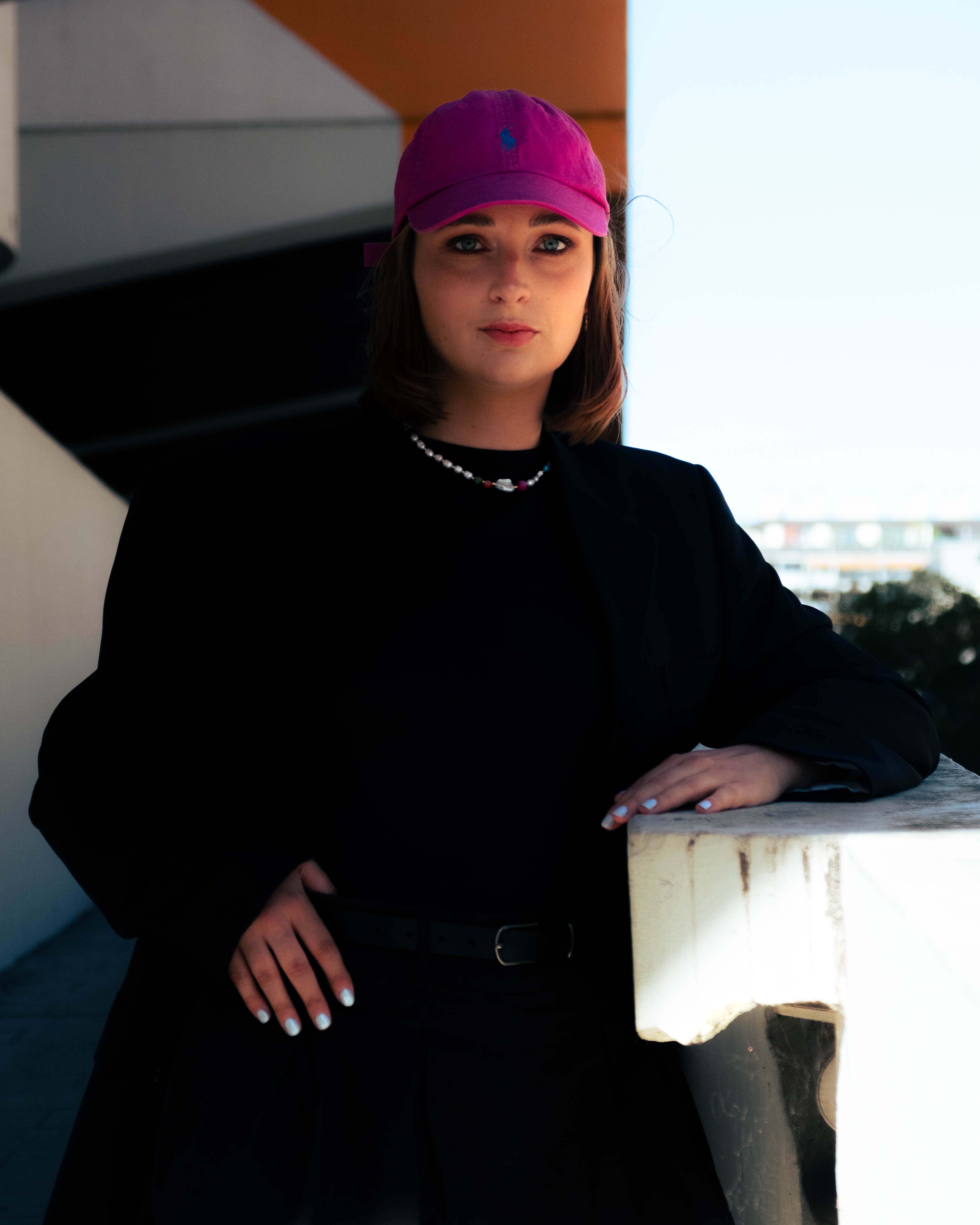 Femme vêtue de noir et portant un collier Bourjeone en perles blanches et colorées, avec une casquette fuchsia – collection Vitaminée.