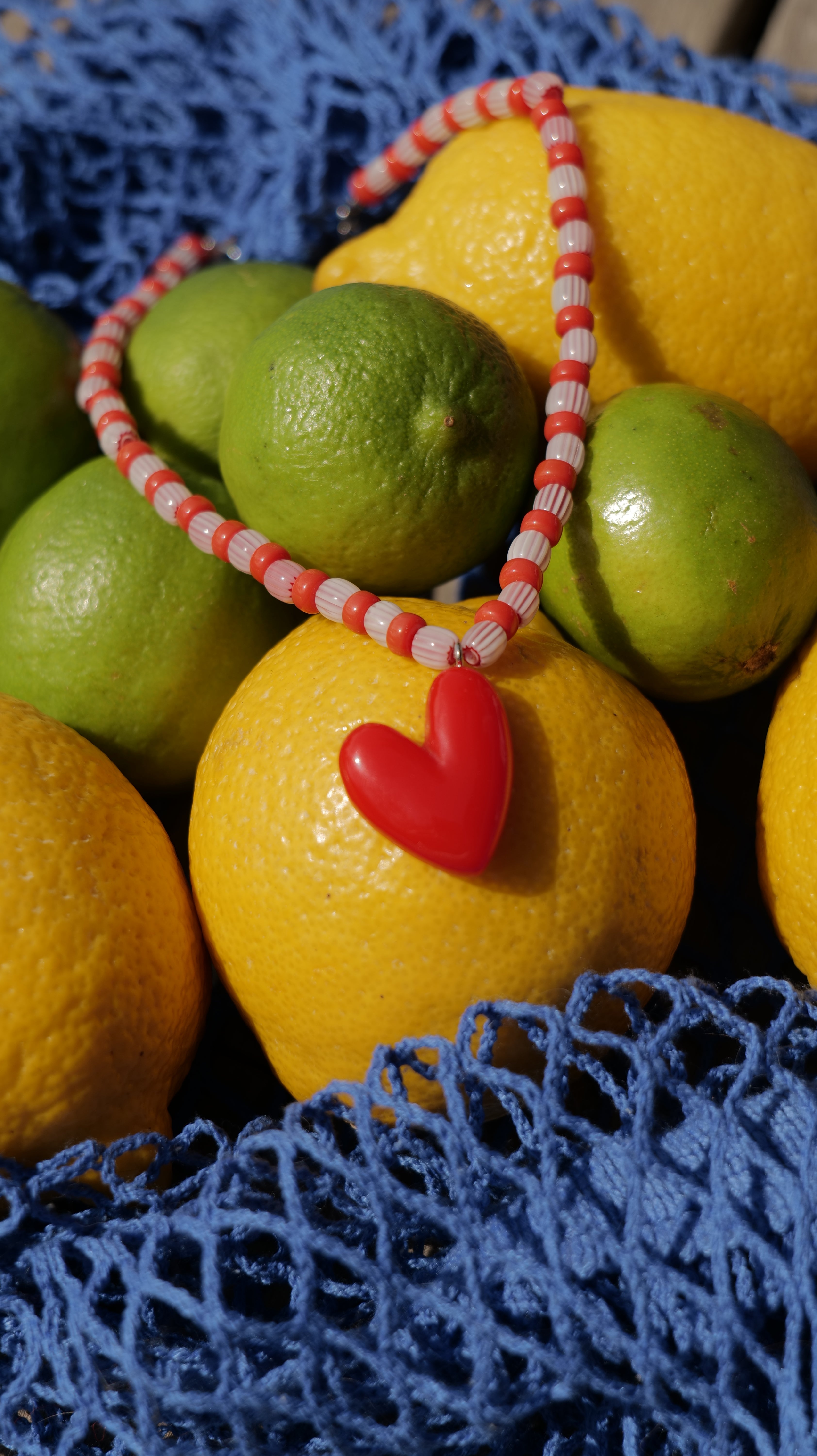 Collier Bourjeone rouge et blanc en perles avec un pendentif cœur, photographié sur des citrons et des limes – collection Umami.