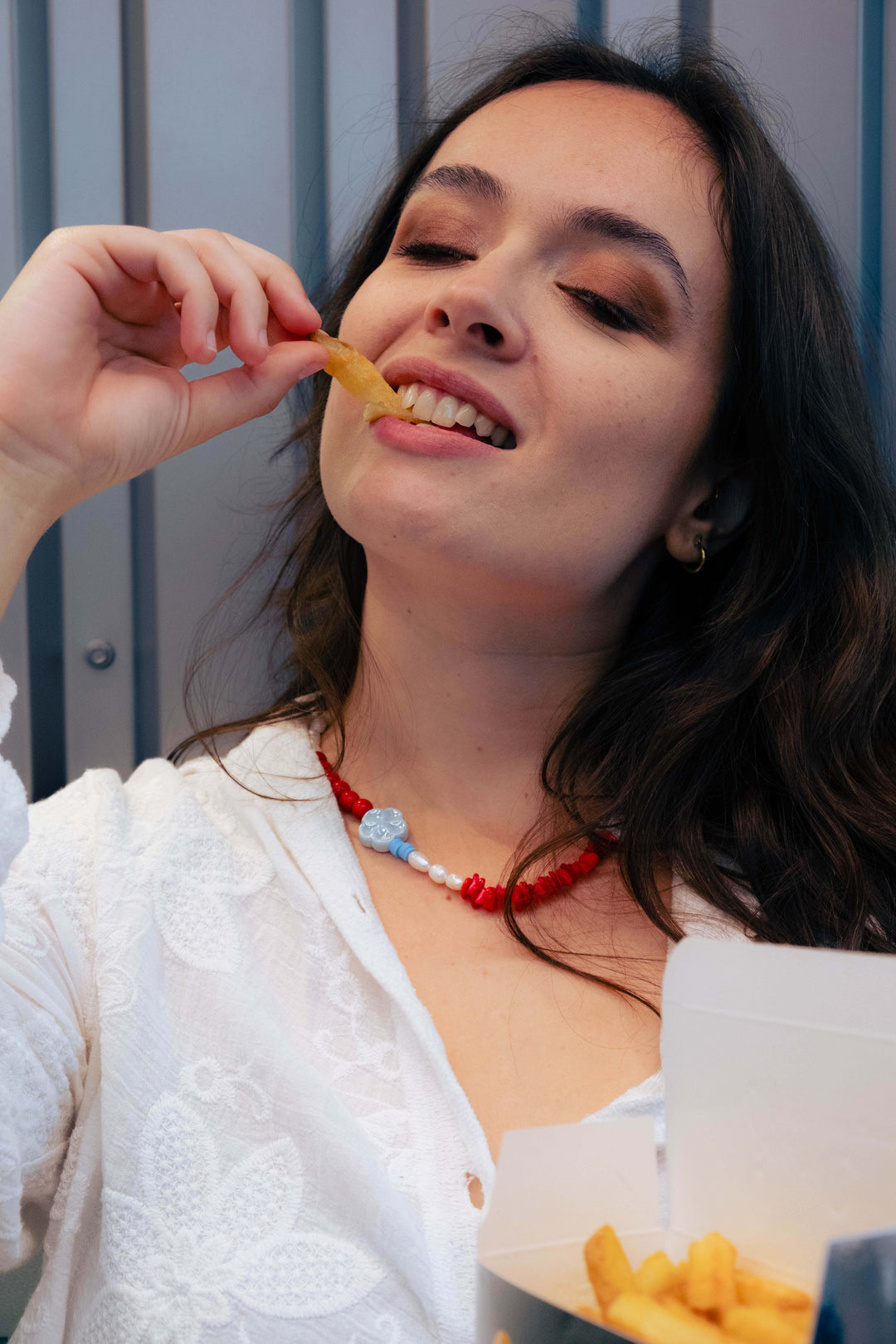 Femme portant un collier Bourjeone rouge et bleu en perles artisanales, dégustant des frites avec sensualité – collection Umami.