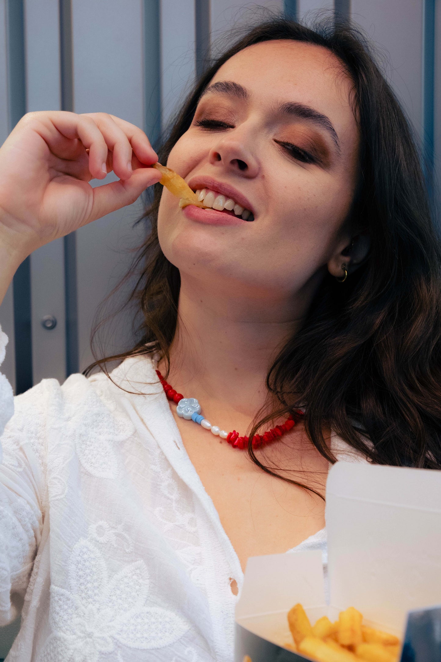 Femme portant un collier Bourjeone rouge et bleu en perles artisanales, dégustant des frites avec sensualité – collection Umami.