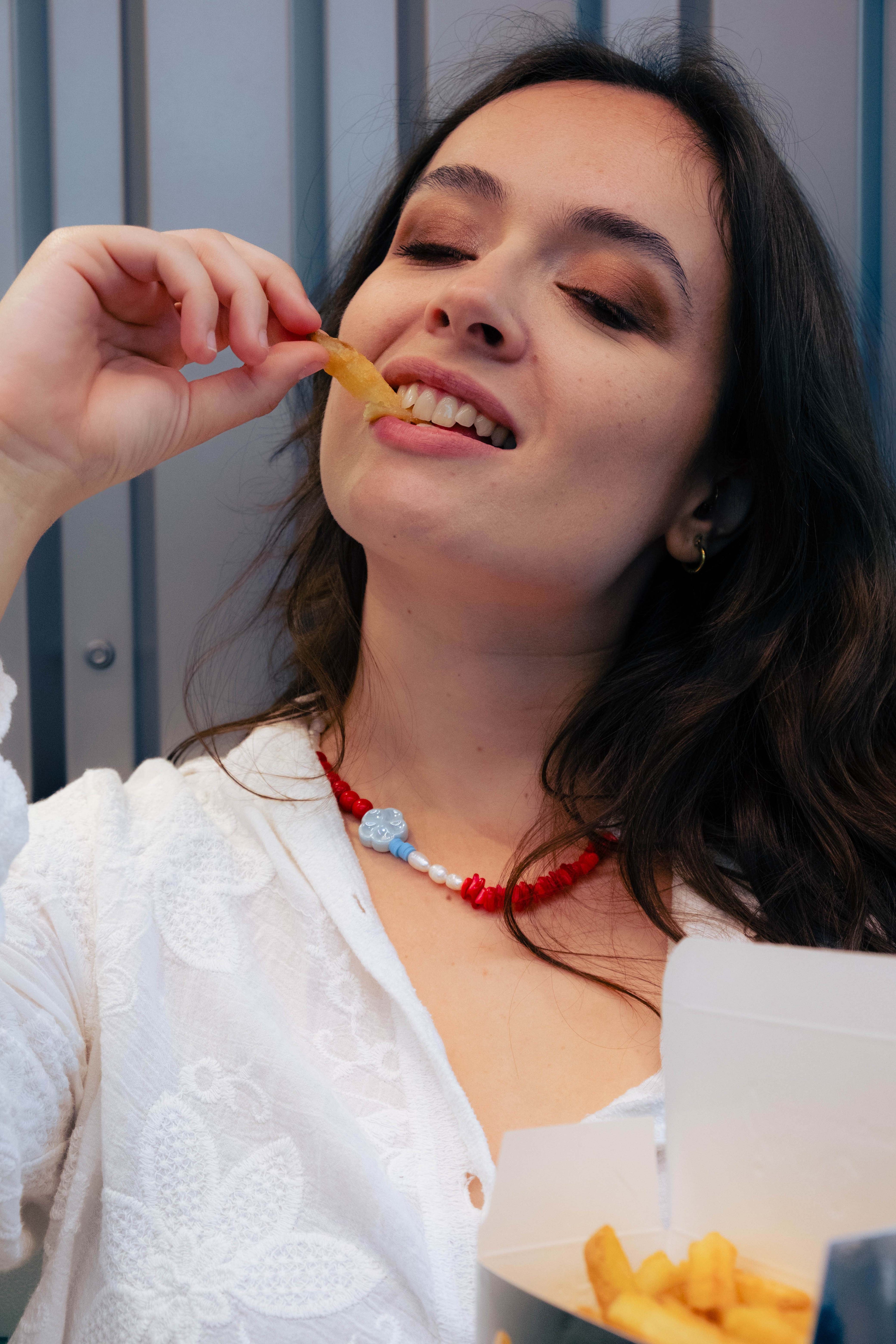 Femme portant un collier Bourjeone rouge et bleu en perles artisanales, dégustant des frites avec sensualité – collection Umami.