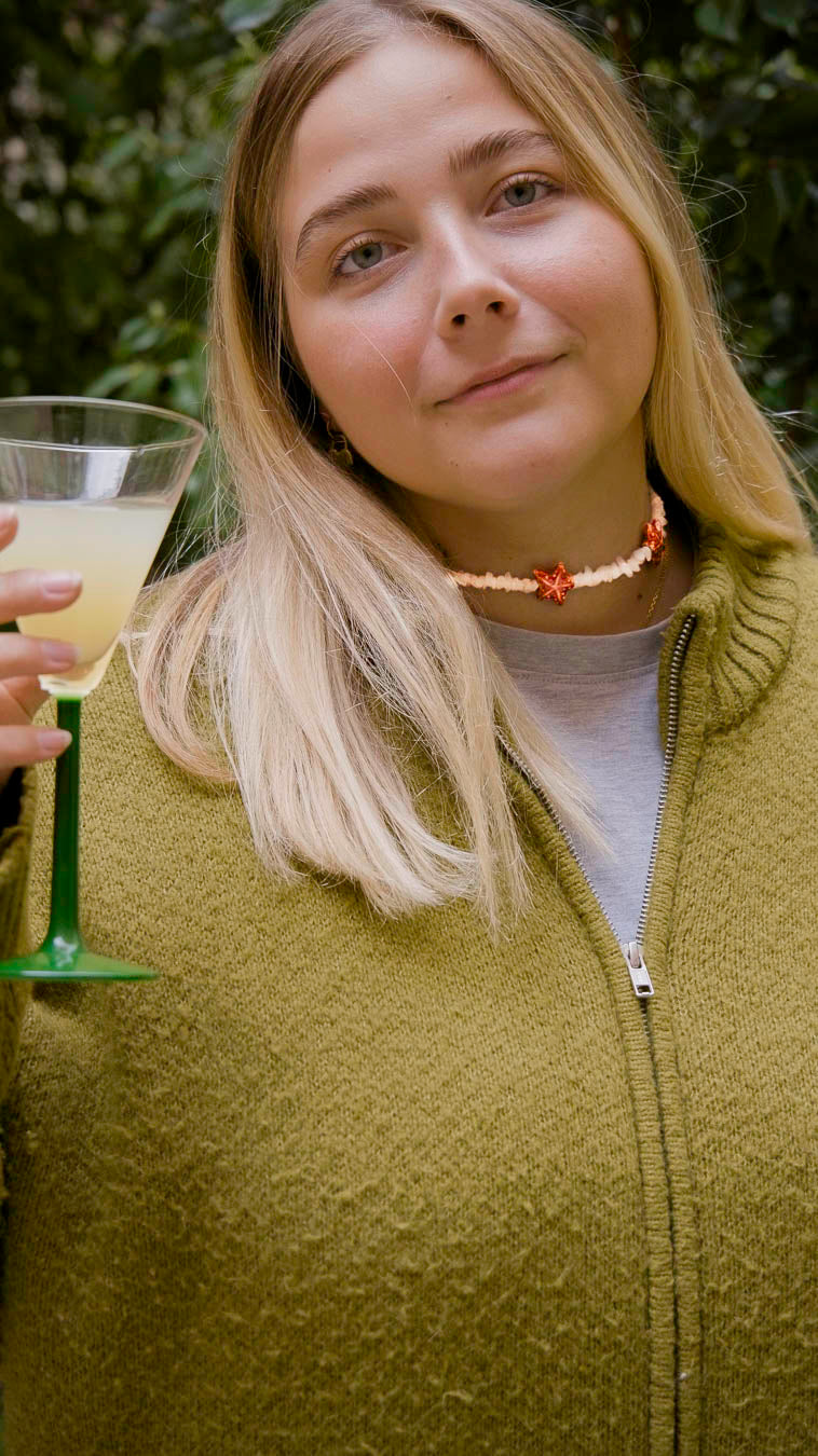 Femme portant un collier Bourjeone orné de coquillages et d’étoiles orangées, levant un verre dans un jardin.