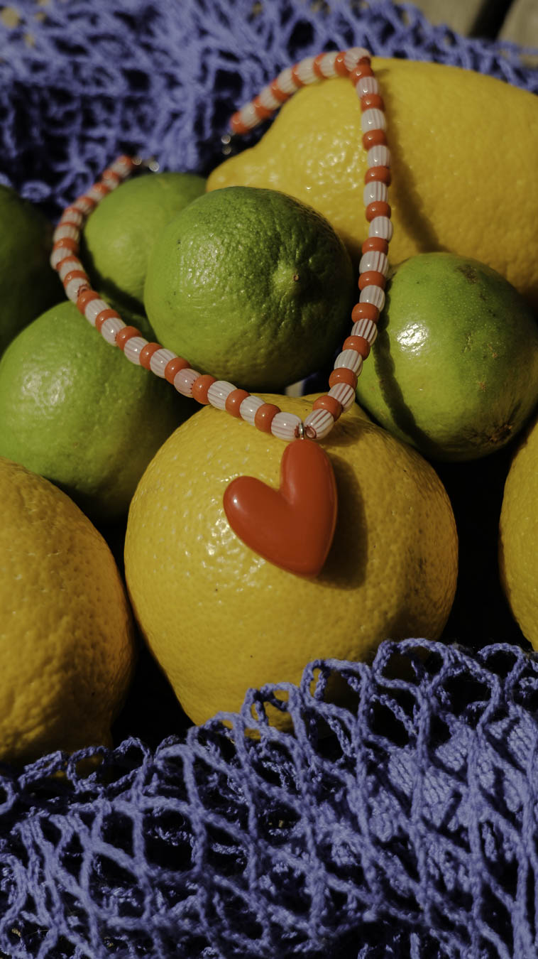 Collier Sucre d’Orge Bourjeone posé sur des citrons et citrons verts, bijou fait main en perles rouges et blanches avec pendentif cœur, doux et rétro.