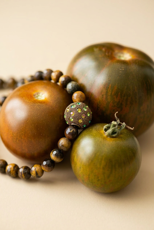 Gros plan du collier Gravy unisex, mêlant perles facettées en œil de tigre brun et une perle centrale ornée de strass multicolores, posé audacieusement à côté de tomates noires.