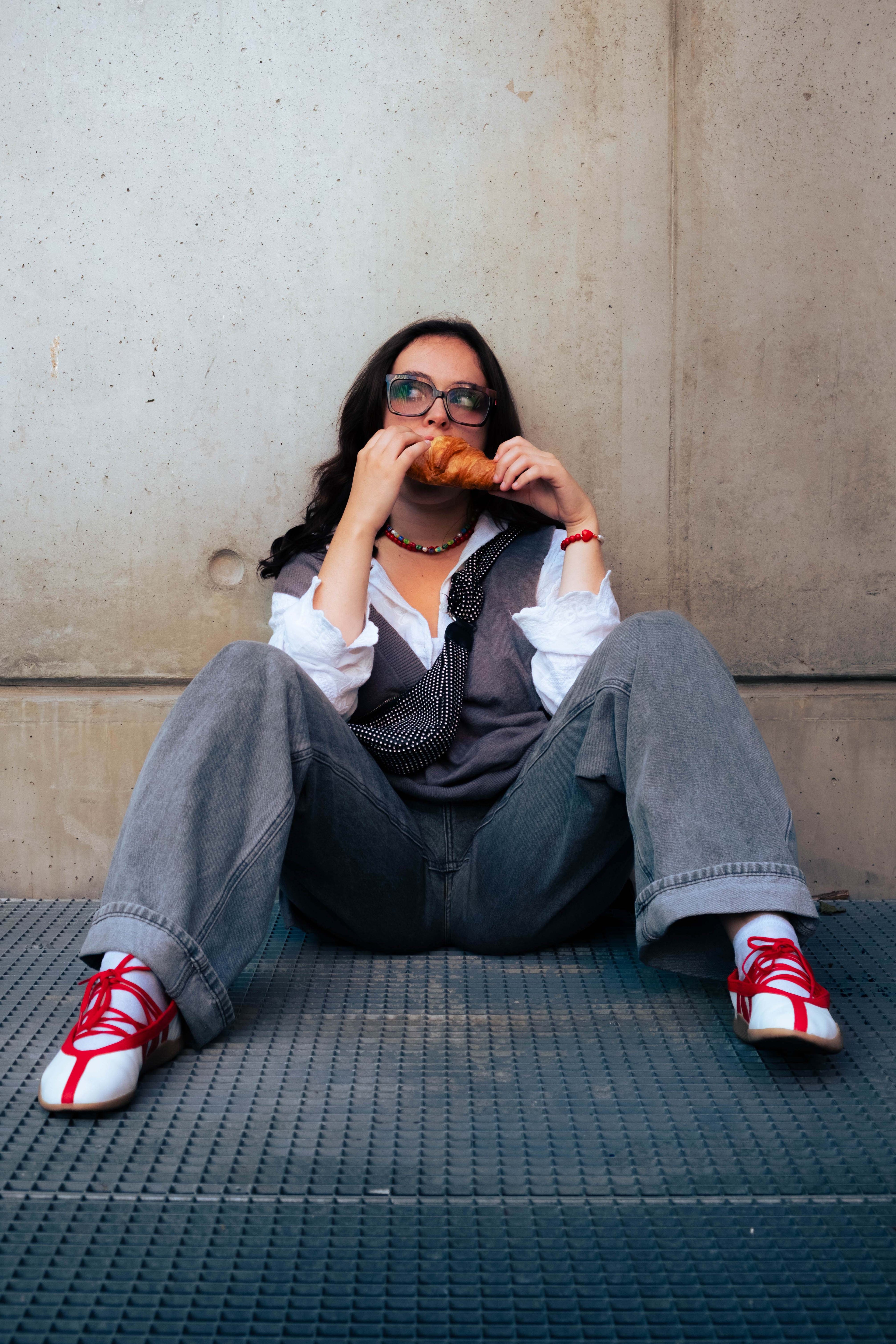 Femme assise contre un mur en jeans gris, portant un collier coloré et un bracelet rouge Bourjeone, dégustant un croissant – collection Umami.
